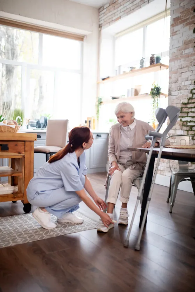 24 Stunden Pflege zu Hause: Pflegerin hilft einer Seniorin im Wohnzimmer